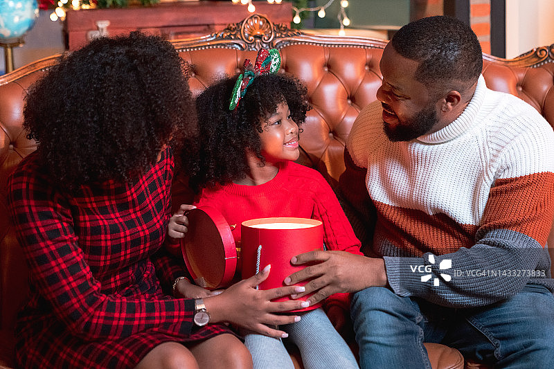 非裔美國小女孩在圣誕前夜和父母坐在裝飾好的客廳的地板上打開禮盒圖片素材