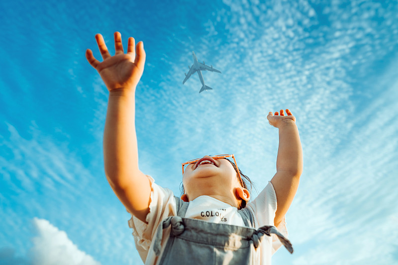 快樂的亞洲小女孩戴著花形太陽鏡開心地微笑著，在湛藍的天空中舉起雙手向飛機揮手圖片素材