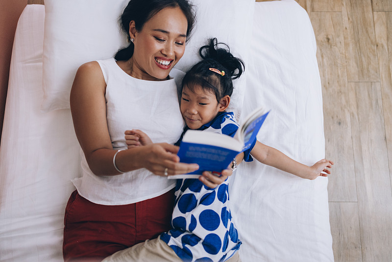 快乐的泰国妈妈躺在床上和她可爱的小女儿和读她的故事前午睡图片下载