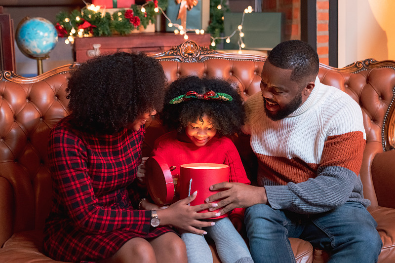 非裔美國小女孩在圣誕前夜和父母坐在裝飾好的客廳的地板上打開禮盒攝影圖片