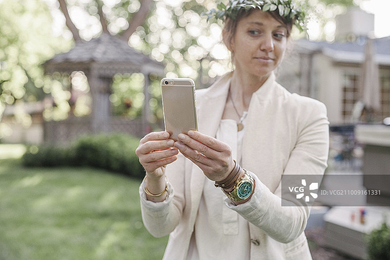 一个头上戴着花圈的女人，站在花园里，用手机自拍。图片素材