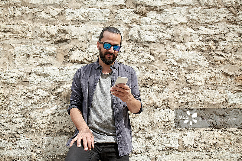 休闲，科技，交流和人的概念-微笑的男人在城市街道的石墙上发智能手机短信图片素材