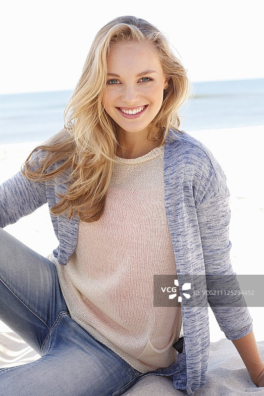一个年轻的金发女郎在海滩上穿着一件浅色针织套衫，一件开襟羊毛衫和牛仔裤图片素材
