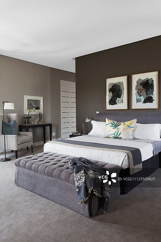 一间雅致的卧室，双人床脚边的棕色搁脚凳，深棕色墙上挂着画框图片素材