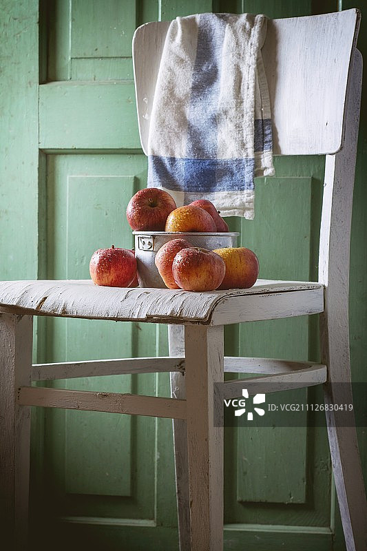 绿色的木墙边，一张旧的白色木椅上放着一碗盛着红苹果的铝碗图片素材