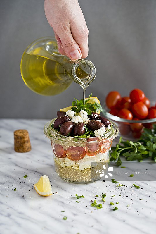 将橄榄油倒在一个玻璃罐中的藜麦沙拉上，配以羊乳酪、番茄和卡拉马塔橄榄图片素材