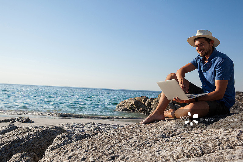 微笑的男人坐在岩石上，在海滩上使用笔记本电脑图片素材