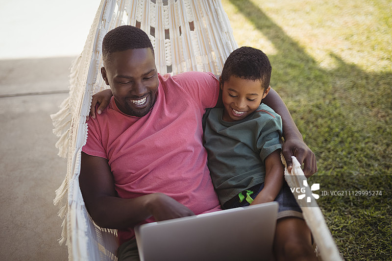 快乐的父亲和儿子在吊床上休息时使用笔记本电脑图片素材