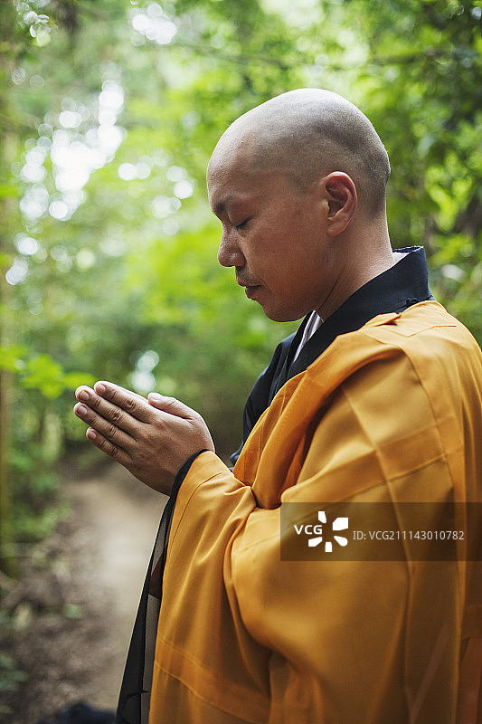 侧面的佛教僧人剃着光头，穿着黑黄僧袍，站在户外，冥想。图片素材