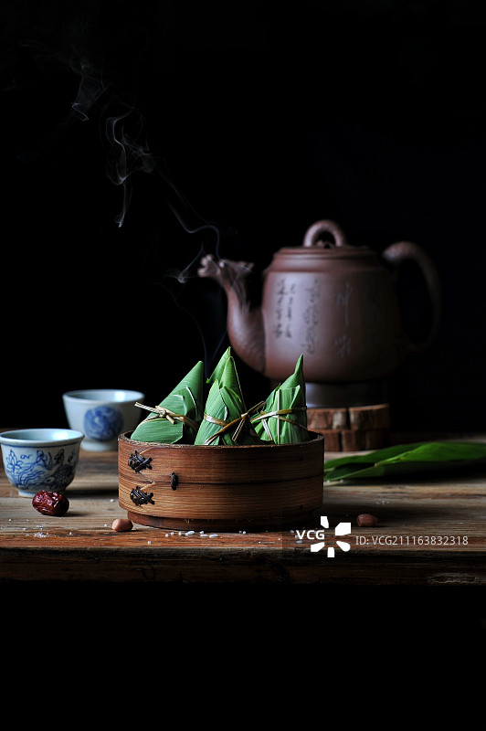 一蒸笼青叶粽子放在冒着水蒸气的紫砂龙壶前，旁边摆着两个青花茶图片素材