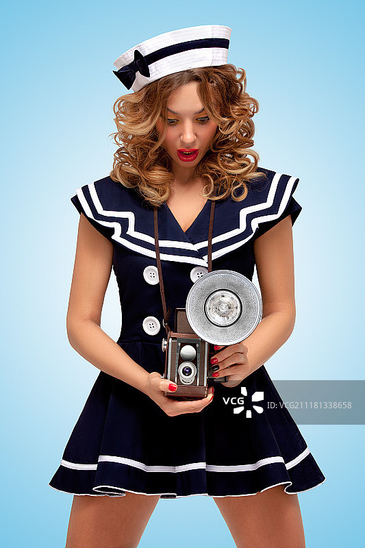 复古照片的一个时尚的海报上的水手女孩看着一个古老的照片相机与灯泡闪光的蓝色背景令人惊讶。图片素材