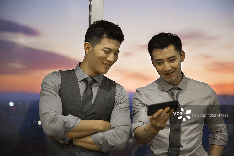 两个青年男商务人士站在办公大厦玻璃窗前看手机沟通工作图片素材