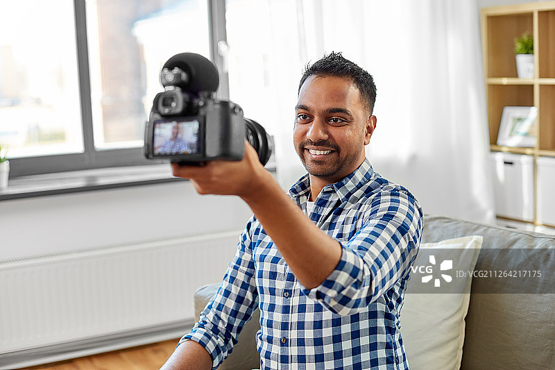 博客，视频博客和人们的概念-微笑的印度男性视频博客与相机视频博客在家里。男性视频博主用相机在家写博客图片素材