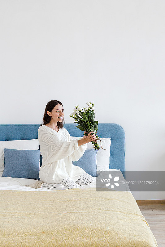 一个穿着白色毛衣的女人坐在床上，手里拿着一束白玫瑰图片素材
