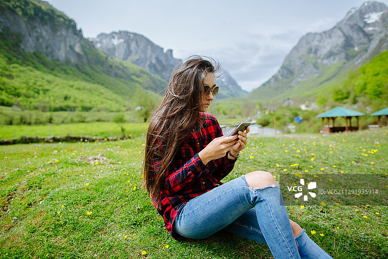 近距离观看的年轻女子与长，棕色头发坐在草地上使用她的电话与凉亭和山的背景，Plav, Plav，黑山图片素材