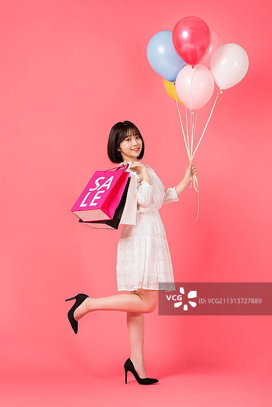 一名年轻女子站在粉红色的背景上，手里拿着购物袋和气球图片素材