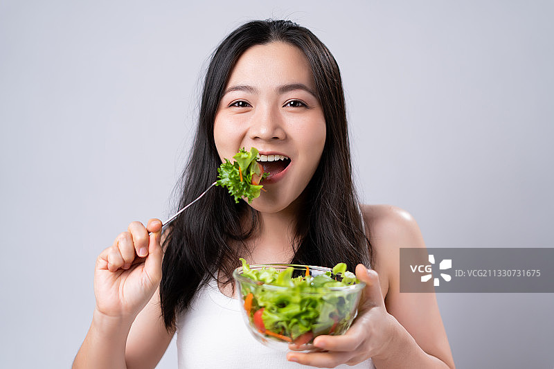 在白色的背景下，微笑的年轻女子吃蔬菜沙拉的肖像图片素材
