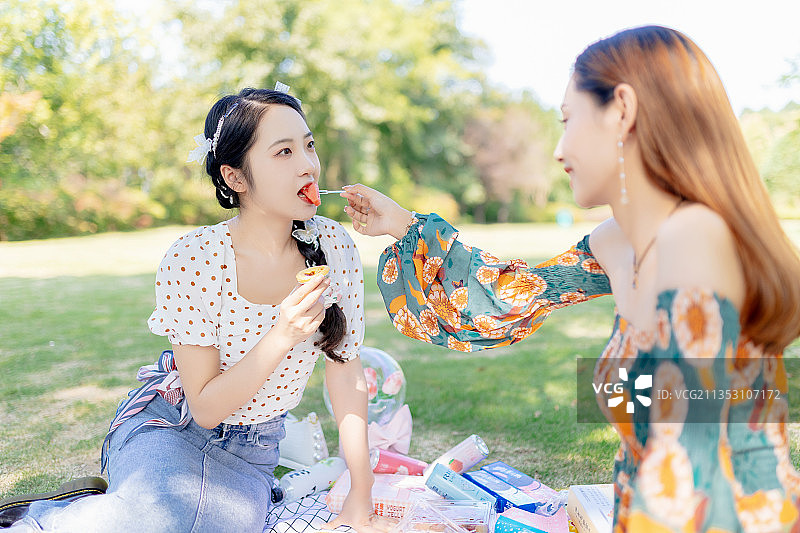 两个亚洲青年美女闺蜜在户外野餐垫上野餐喂东西吃图片素材