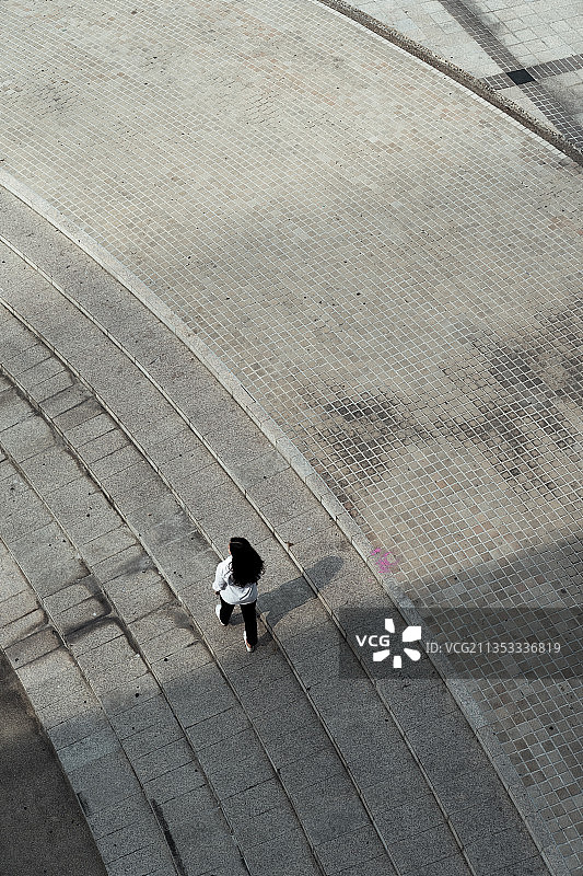 俯视图的黑色长发，穿着白色衬衫的女人走在鹅卵石地板上的广场。图片素材