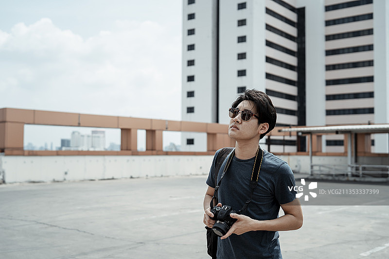 年轻英俊的亚洲摄影师男子戴着太阳镜，拿着相机走在屋顶上寻找灵感。图片素材
