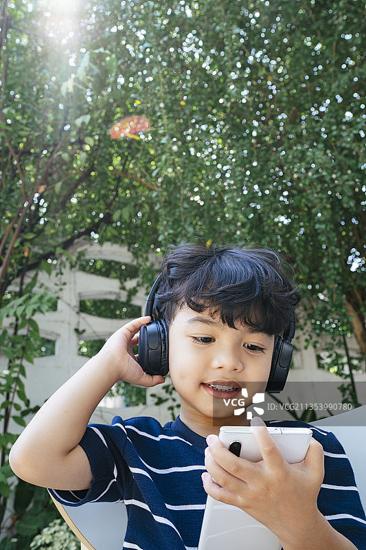 周末，小男孩戴着无线耳机坐在后院，用智能手机看卡通片。图片素材