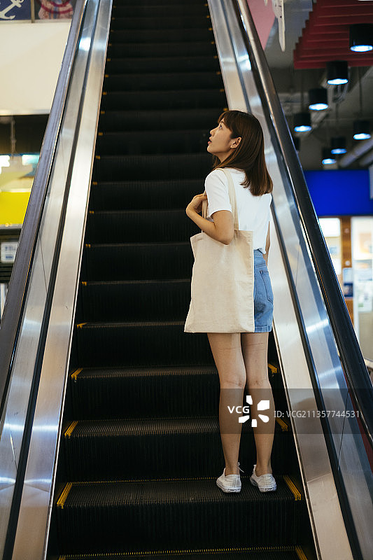 年轻的亚洲泰国女人穿着白衬衫站在自动扶梯上，拿着布质购物袋。图片素材