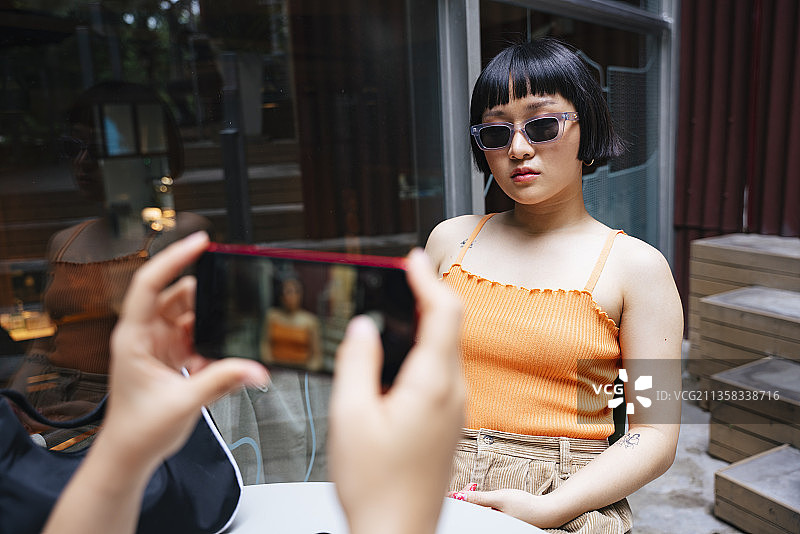 亚洲年轻女性戴着太阳镜，在咖啡馆里被朋友用智能手机拍照。图片素材