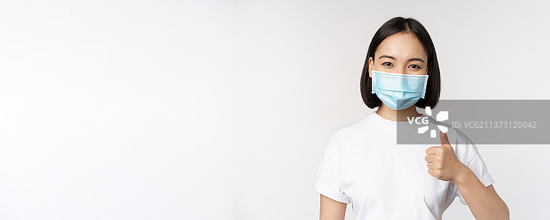 带着医用口罩的亚洲女孩微笑着竖起大拇指，批准图片素材