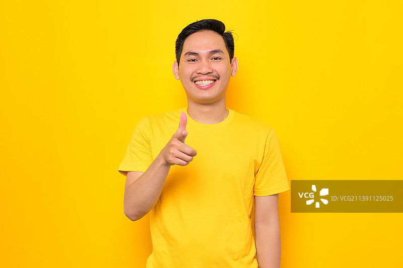 微笑的亚洲年轻男子指着相机孤立的黄色背景，印度尼西亚图片素材
