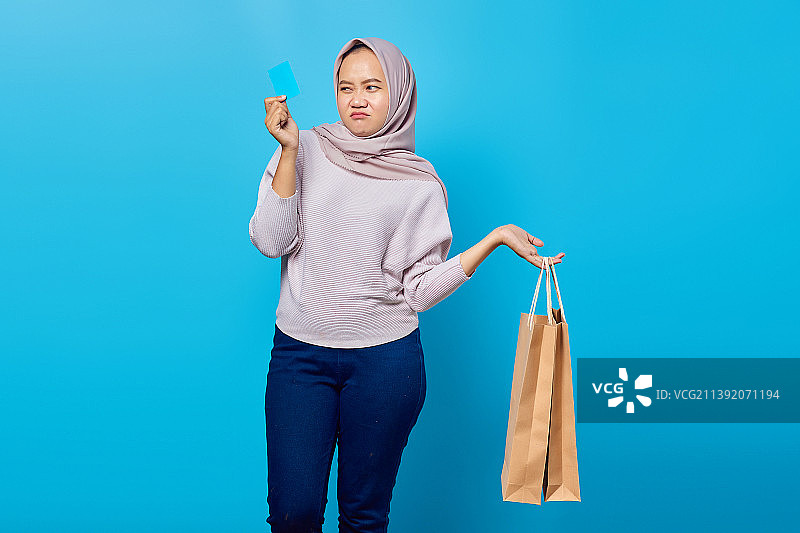 肖像有吸引力的亚洲妇女拿着购物袋和显示信用卡在蓝色背景，印度尼西亚图片素材