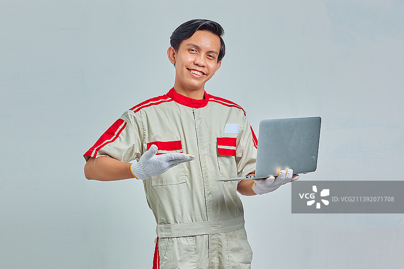 欢快英俊的年轻机械师穿着制服，指着灰色背景的笔记本电脑，印尼图片素材