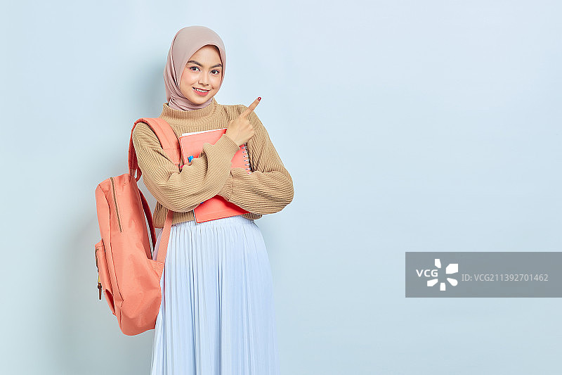 微笑的年轻亚洲穆斯林女学生在棕色毛衣，指着一边的复印空间图片素材