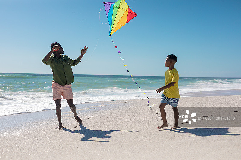全长度的非洲裔美国父亲和儿子跑与风筝在海滩上，对着晴朗的蓝天图片素材