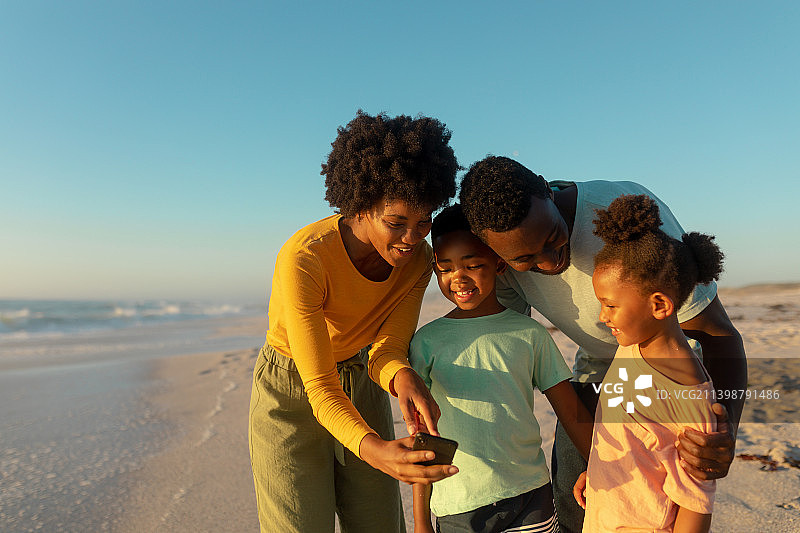 阳光明媚的日子，快乐的非裔美国母亲在海滩上与家人分享智能手机图片素材