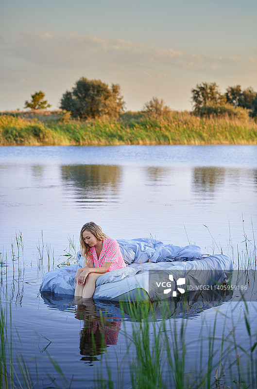 一个穿着粉红色圆点睡衣的严肃的金发女人坐在湖中央的床上。很适合做书的封面。超现实主义的概念图片素材