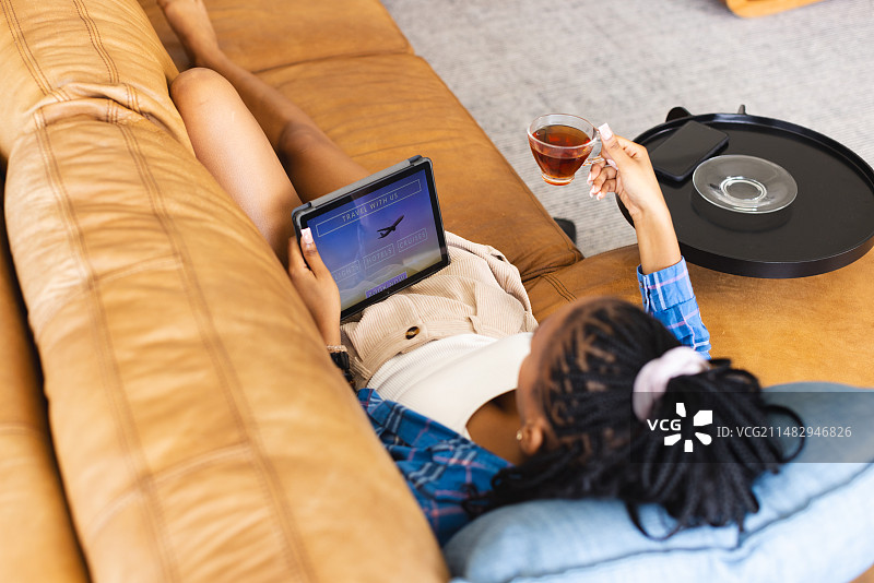 一位年轻的非裔美国女子躺在沙发上，手里拿着平板电脑和饮料，在网上预订假期图片素材