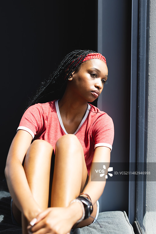 一位年轻的非裔美国妇女坐在窗边，阳光照亮了她若有所思的表情图片素材