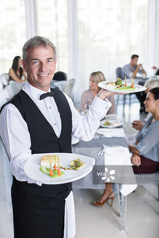 微笑的服务员拿着花哨的盘子的肖像，人们坐在餐厅的桌子在背景图片素材