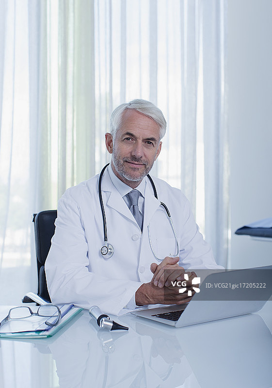 微笑的成熟医生坐在办公桌上的笔记本电脑在办公室图片素材