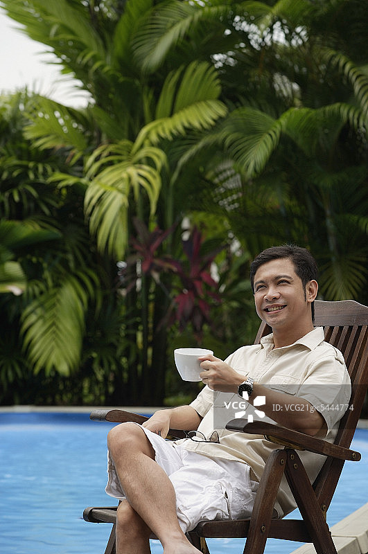 一个男人坐在游泳池旁，拿着一杯咖啡，微笑着图片素材
