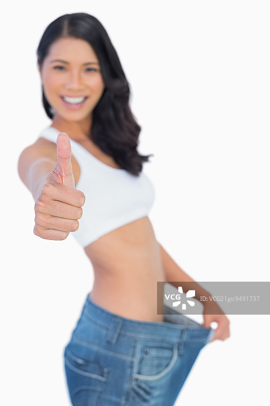 胜利的女人拿着她太大的裤子大拇指对着白色的背景图片素材