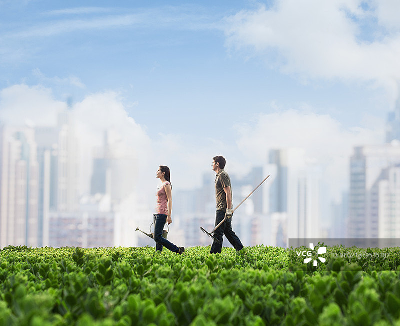 两个年轻人扛着园艺设备穿过一片绿油油的田野，背景是城市景观图片素材