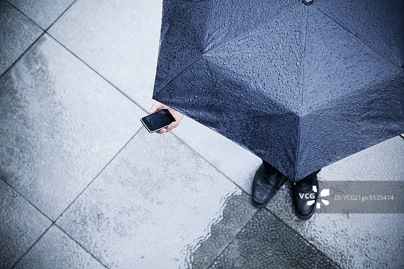 高视角的商人拿着伞，看着他的手机在雨中图片素材