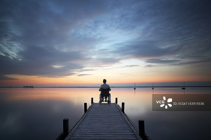 夕阳西下时，一个老人坐在码头上的轮椅上图片素材