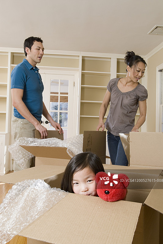 女孩(4-5岁)从客厅的纸箱里探出头来，父母在后面工作图片素材