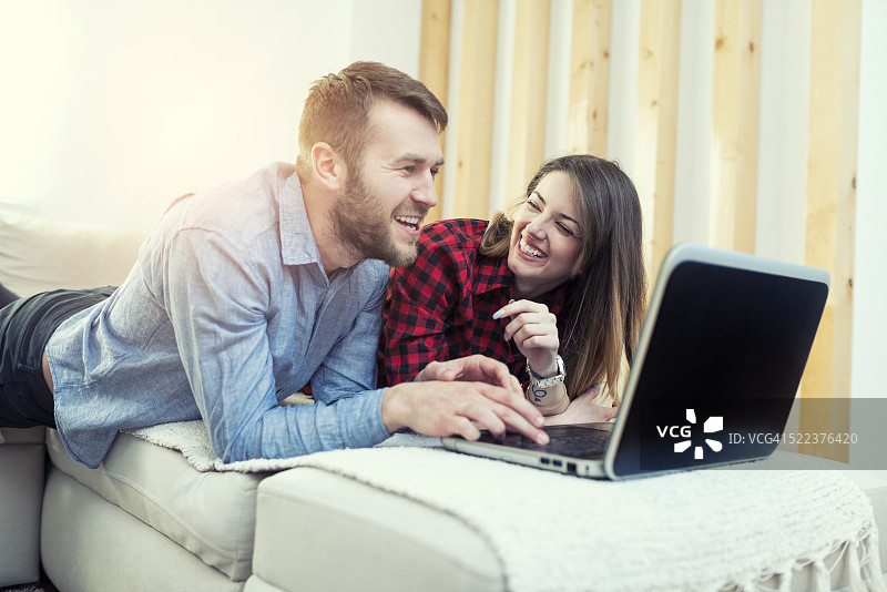 微笑的夫妇躺在沙发上，在家里使用笔记本电脑图片素材