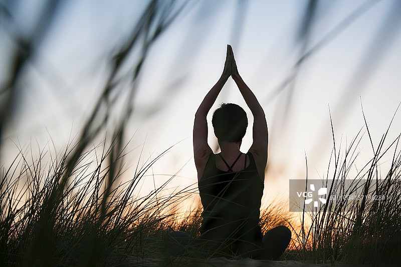 中年成年妇女在日落时在剪影长草练习瑜伽的后视图图片素材