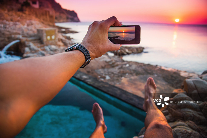 夏日里，在地中海科斯塔布拉瓦海岸线的一个天然泳池里，一名男子用智能手机拍下了自己的照片，他的双腿漂浮在空中，观看日出。图片素材