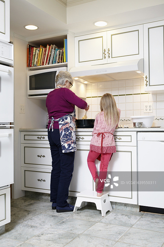 后视图的女孩和奶奶搅拌锅在厨房的铁架上图片素材