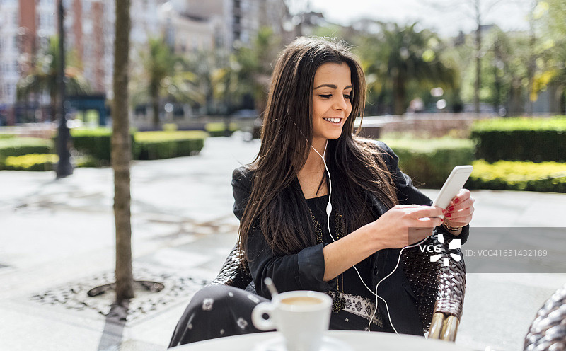 年轻漂亮的女人在喝咖啡，路边咖啡馆，玩智能手机，听音乐图片素材